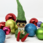 A needle felted elf in a green pointy hat and red pointy shoes sits amongst some holiday decorations.