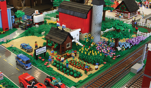 Photo of a LEGO train display with a red barn, gray silo, flower beds, and train tracks.