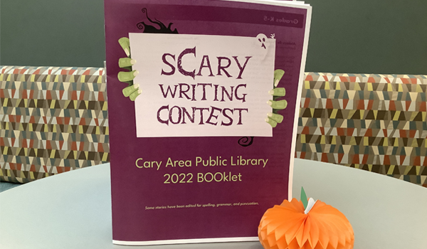 Photo of the sCary Writing Contest BOOklet resting on a tabletop and a small paper pumpkin as decoration.