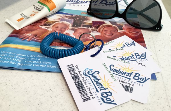 Four Sunburst Bay pool passes on a keyring, a pair of sunglasses, a tube of sunscreen, and the Cary Park District Sunburst Bay brochure displayed on a table.