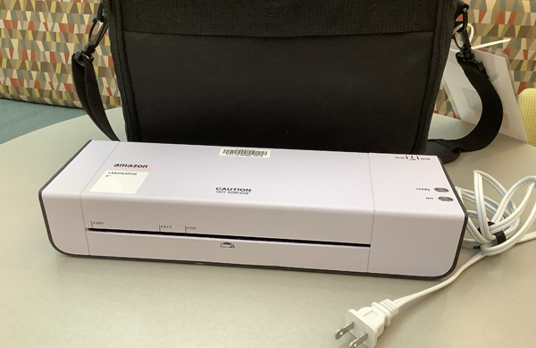 Photo of a white laminating machine with a black case behind it on a tabletop.