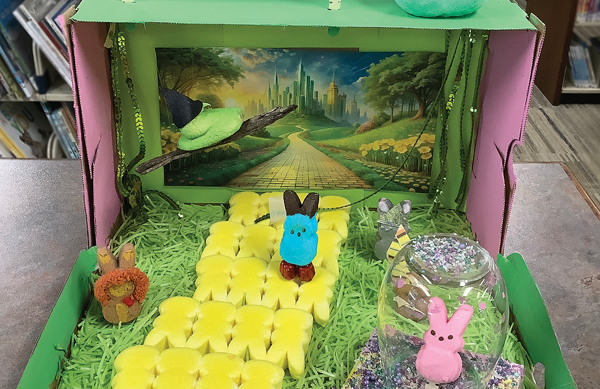 Inside a Wicked Peeps diorama with yellow Peeps forming a yellow brick road, a green Peep as a witch flying, and a pink Peep standing to the side within a glass jar bubble.