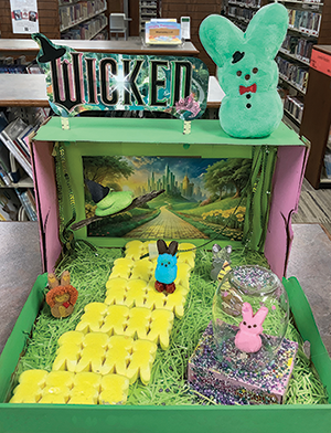 Wicked-themed Peeps diorama with yellow Peeps creating a yellow bring road with a green "witch" Peep flying above and a pink Peep in a glass jar "bubble" along side.