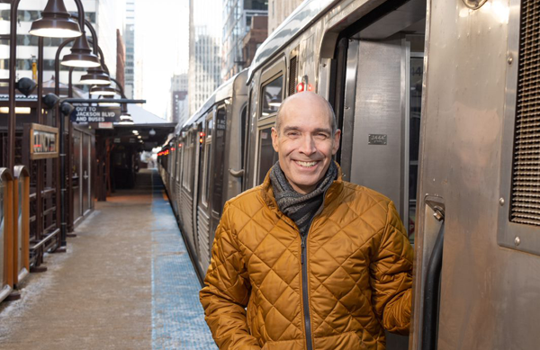 A man in a pale orange coat and gray scarf smiles towards the camera as he steps out of an open subway cart door.