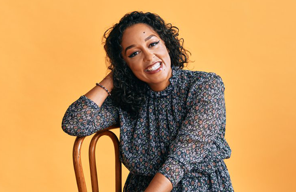 A woman is sitting in a chair with her arm resting on the back and propping up her tilted head and she smiles towards the camera.